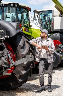 Agricultural equipment dealer or supplier. Caucasian man in plaid shirt stands next to tractor and using digital tablet.	