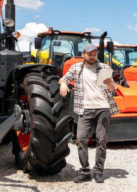 Agricultural equipment dealer or supplier. Caucasian man in plaid shirt stands next to tractor and using digital tablet.	