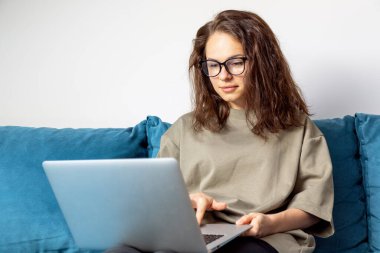 Beyaz kadın gözlük takıyor. Evdeki mavi koltukta otururken laptop kullanıyor..