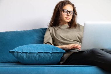 Beyaz kadın gözlük takıyor. Evdeki mavi koltukta otururken laptop kullanıyor..