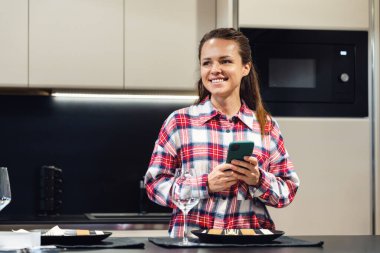 Gülümseyen kadın elinde cep telefonuyla mutfakta duruyor.