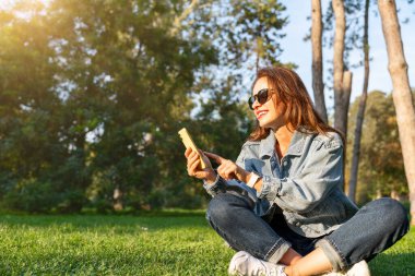 Şehir kadını akıllı telefonunu kontrol ediyor ve yazın çimenlikte otururken gülümsüyor.