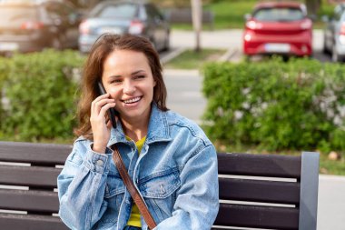 Gündelik kıyafetli genç bir kadın sokak bankında oturmuş cep telefonuyla konuşuyor.