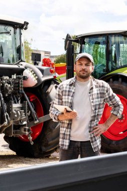 Erkek çiftçi, teçhizat bayiindeki şöhret traktörlerinin arka planında dijital tablet tutuyor.