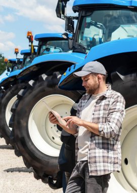 Erkek çiftçi, makine bayisinde dijital tablet kullanarak traktör tekerleğine yaslanıyor.