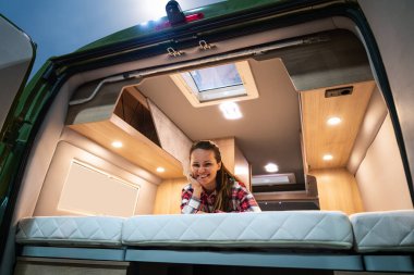 Karavanda yatakta dinlenen bir kadın kameraya bakıyor ve gülümsüyor. Karavan yaşam biçimi.