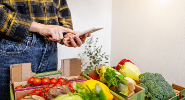 Genç kadın çiftçi online siparişe hazırlanıyor akıllı telefon kullanarak organik sebzeleri teslimat kutularında paketliyor.