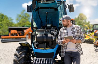 Erkek çiftçi, modern teknolojiyle traktör motorunu incelemek için dijital tablet kullanıyor. Tarım makineleri tanısı.