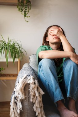 Yorgun bir kadın kanepede oturuyor ve evde elini yüzünü kapatıyor.