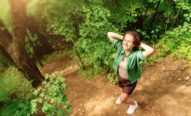 Elleri başının arkasında gülümseyen kadın doğal bir parkta yürümekten zevk alıyor.