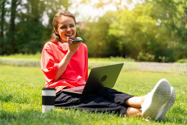 Şehir parkında dizüstü bilgisayarla dinlenirken genç bir kadın sesli mesaj kaydediyor. Dijital iletişim.