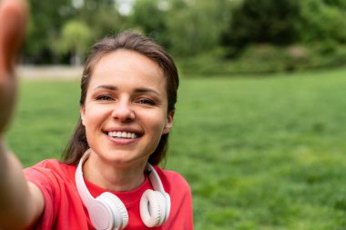 Kırmızı tişörtlü genç kadın şehir parkında kulaklıklı selfie çekiyor.