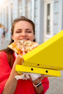 Kırmızı tişörtlü mutlu genç kadın canlı bir yaz gününde açık havada pizza yiyor.