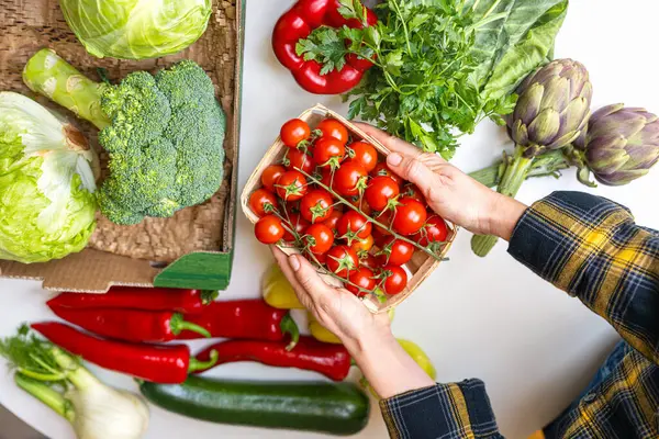 Masanın üzerinde kirazlı domateslerle dolu bir kutuyu tutan eller.