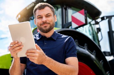 Profesyonel tarım işinin portresi traktörün önünde dijital tablet tutuyor.