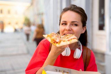 Kırmızı tişörtlü şık bir kadın şehirde pizza yiyor.