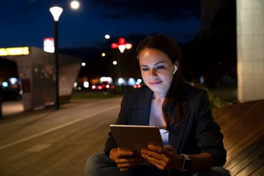 Modern kadın geceleri şehir caddesinde dijital tablet kullanarak bankta oturuyor.