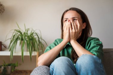 Şok olmuş ve stresli bir kadın. Elleri suratında. Evdeki kanepede oturuyor.