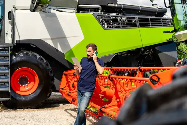 Tarım endüstrisi çalışanı tablet kullanıyor ve traktörün yanında cep telefonuyla konuşuyor.