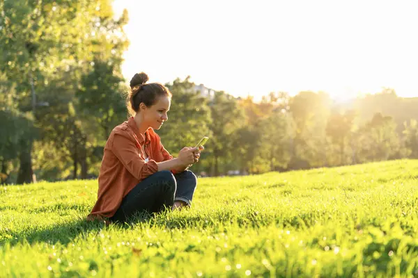 Modern kadın çimlerin üzerinde oturuyor ve akıllı telefonunu kullanarak mesaj yolluyor..