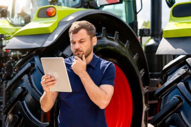 Düşünceli adam traktörün yanındaki dijital tablet hakkında bilgi analiz ediyor.
