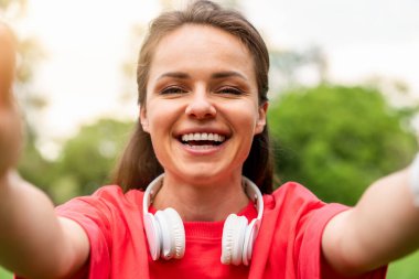 Boynunda kulaklıkları olan neşeli bir kadın dışarıda selfie çekiyor.