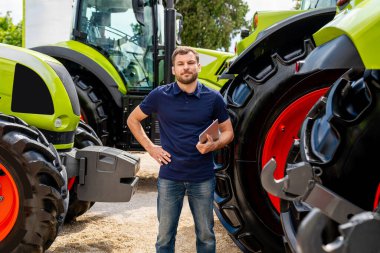 Açık hava tarım bayiliğinde traktörlerin arasında duran tabletli adam.