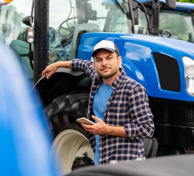 Kendine güvenen erkek çiftçi, tarım ekipmanları bayisinde mavi traktörün yanında akıllı telefon tutuyor.