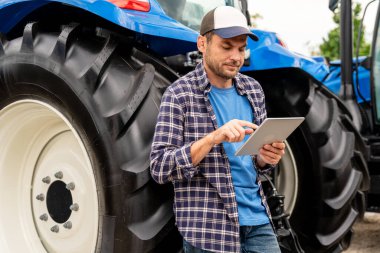 Erkek çiftçi açık havada traktörün yanında dijital tablet kullanarak kep ve ekose gömlek giyiyor..