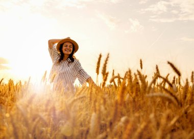 Hasır şapkalı gülümseyen kadın gün batımında mısır gevreği tarlasında şehir kaçışının tadını çıkarıyor.