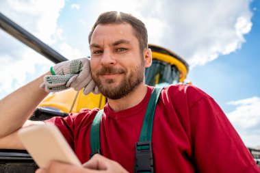 Gülümseyen adam biçerdöver yakınında akıllı telefon kullanarak operatörü birleştiriyor.