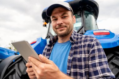 Kapaklı erkek çiftçi dışarıda traktörün önünde dijital tablet tutuyor..