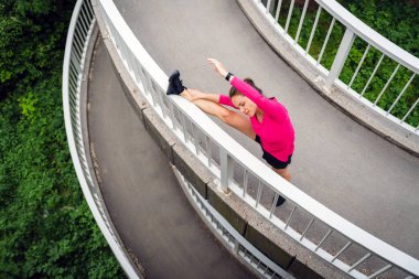 Pembe spor kıyafetleriyle şehir köprüsünde uzanan genç bir kadın.
