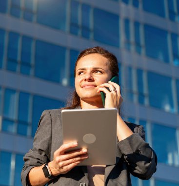 Tablet tutan ve ofis binasının yakınında cep telefonuyla konuşan şehir kadını.