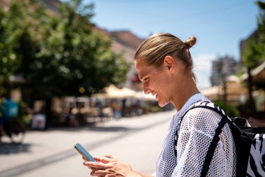 Gülümseyen modern kadın yaz seyahati sırasında şehir caddesi önünde akıllı telefonunu kontrol ediyor.