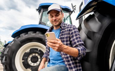 Genç yetişkin erkek çiftçi dışarıda traktörle akıllı telefonunu kontrol ediyor..