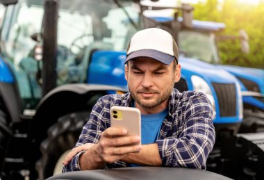 Açık havada traktörlerin önünde günlük kıyafet ve şapka giyen bir çiftçi akıllı telefonu kontrol ediyor.