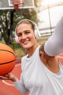 Kablosuz kulaklık takan, elinde basketbol topu tutan ve akıllı telefonuyla selfie çeken genç şehirli kadın.