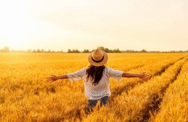 Sıcak güneş ışığı altında kırsal kesimlerde buğday tarlalarıyla çevrili sıradan giysili ve hasır şapkalı bir kadın.