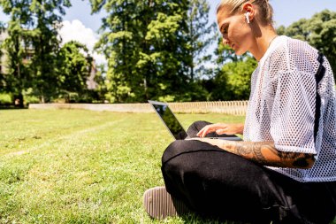 Parkta çimlerin üzerinde otururken dizüstü bilgisayarda çalışan serbest çalışan bir kadın..
