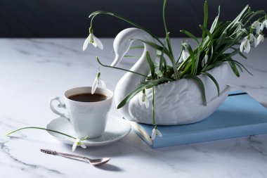 Still life with white snowdrops in a ceramic vase in the form of a swan, coffee in a white cup, conceptual morning ritual whith a book.High quality photo