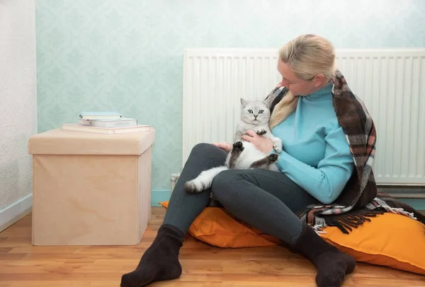 Woman sitting with cat under blanket warming near an electric heater, Cat makes blonde lady happy sitting near heating radiator High quality photo