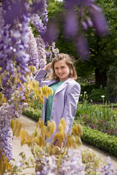 A young woman in a lilac jacket poses near the flowering wisteria in the Botanical Garden. High quality photo