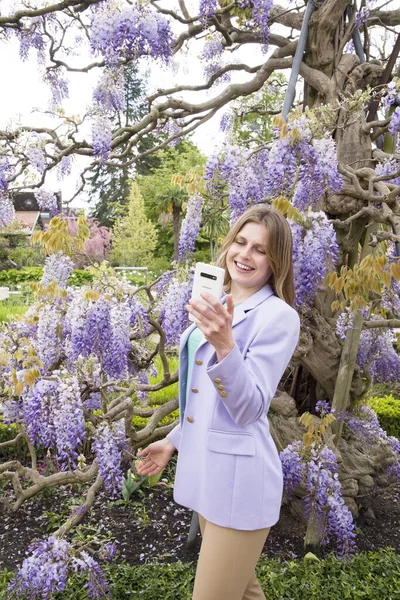 A young woman in a lilac jacket makes selfie with the flowering wisteria in the Garden, Using technology, High quality photo