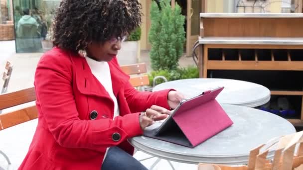 一位身穿红色外套的年轻的非洲裔美国妇女坐在咖啡店的桌子旁 用平板电脑购物后检查帐单 财务问题 开支的合理计划 高质量的镜头 — 图库视频影像