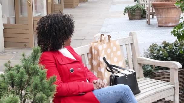 Μια Νεαρή Αφροαμερικάνα Κόκκινο Παλτό Κάθεται Ένα Παγκάκι Μετά Ψώνια — Αρχείο Βίντεο