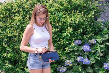 Genç bir kadın küçük çantasında bir şey arıyor. Güzel elbiseler, aksesuarlar, kaliteli fotoğraflar.