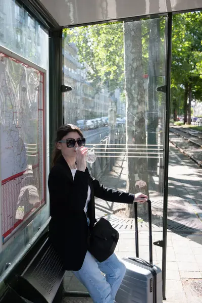 サングラスのビジネス女性が街のトラム停留所で水を飲み黒のエレガントなジャケットを着てスーツケースを手に — ストック写真