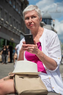 60 yaşında, beyaz şapkalı ve şık yaz kıyafetleri giyen beyaz bir kadın şehirdeki bir bankta akıllı bir telefonla sohbet ediyor sırt çantası yakınlarda duruyor, kaliteli bir fotoğraf.