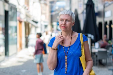 Şık giyimli orta yaşlı bir kadın kentte güvenle gülümsüyor, 60 yaşında mavi bluzlu, gri saçlı bir bayan ve sarı çantalı, yoldan geçenlerin ve dükkanların bulunduğu bulanık bir cadde.